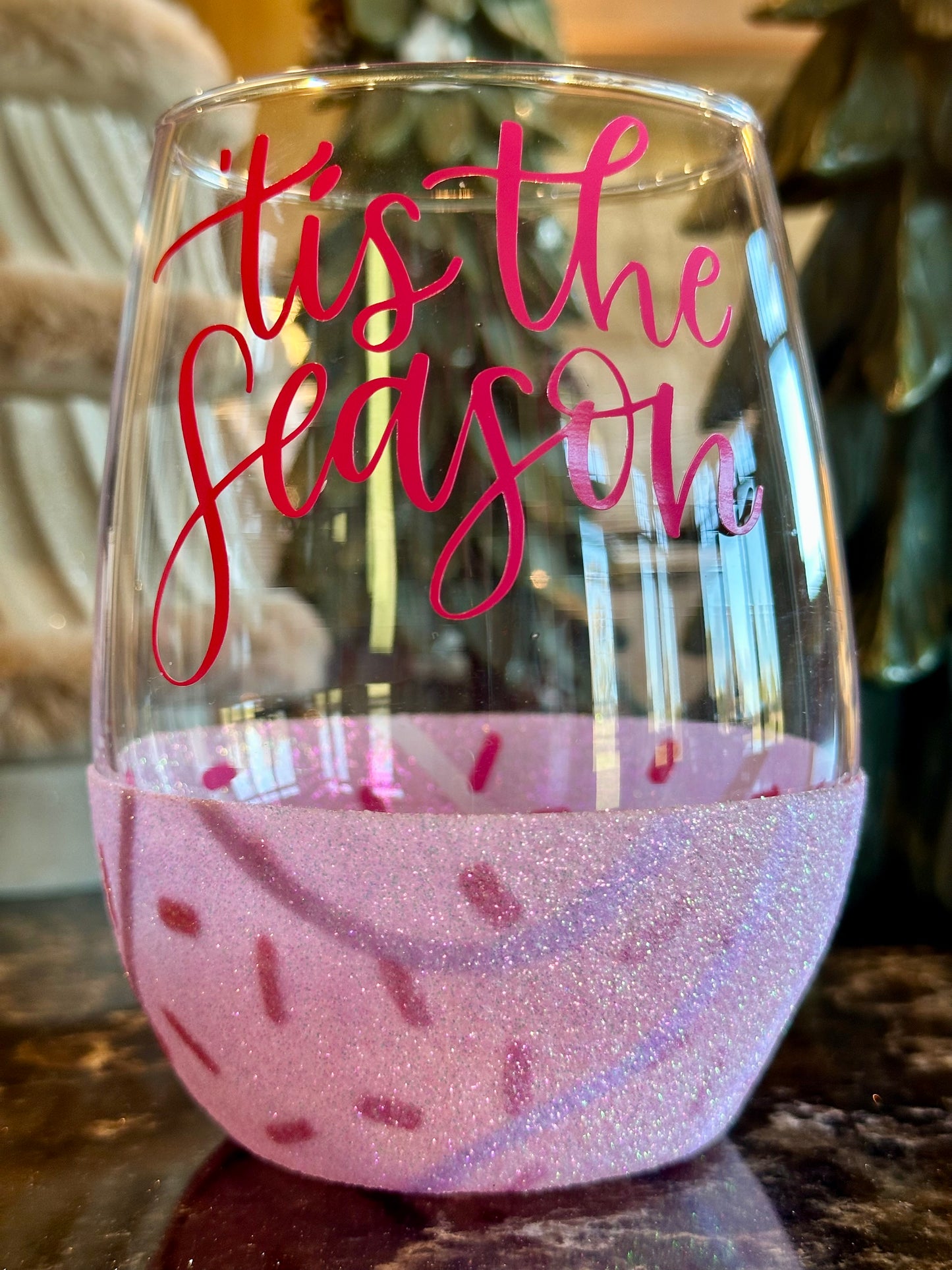 Wine glass with 'tis the season text and pink glitter base on a blurred background