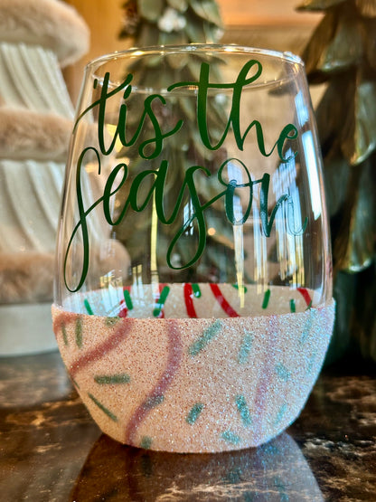 Clear wine glass with 'tis the season' text and glittery base on a reflective surface.